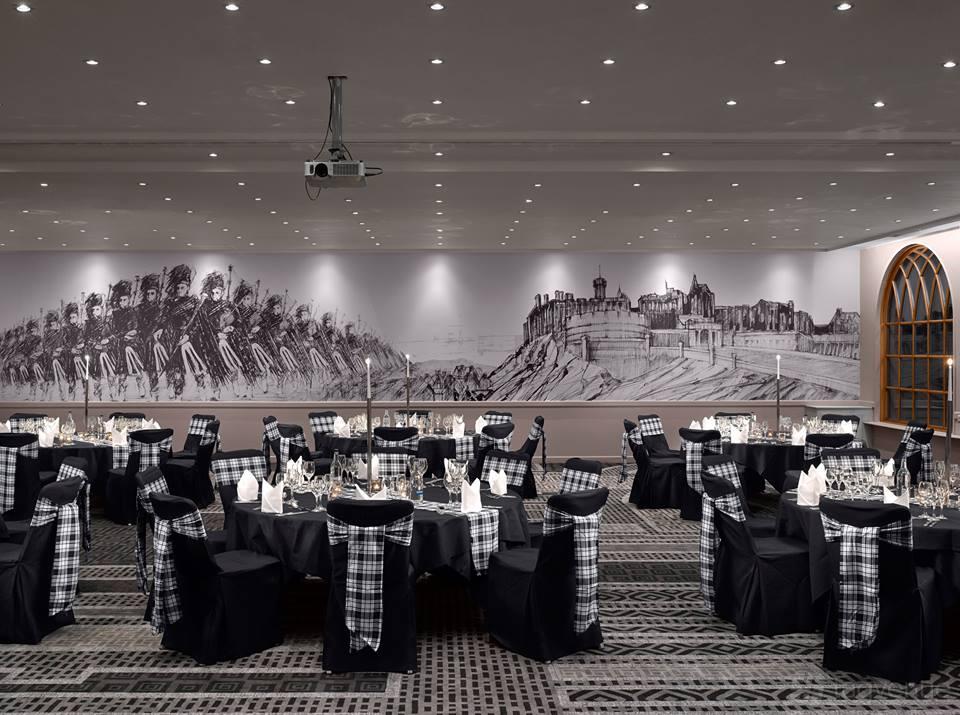 An event space in a hotel with round tables, black chair covers, tartan sashes, and a mural of Edinburgh Castle at Radisson Blu Hotel, Edinburgh City.