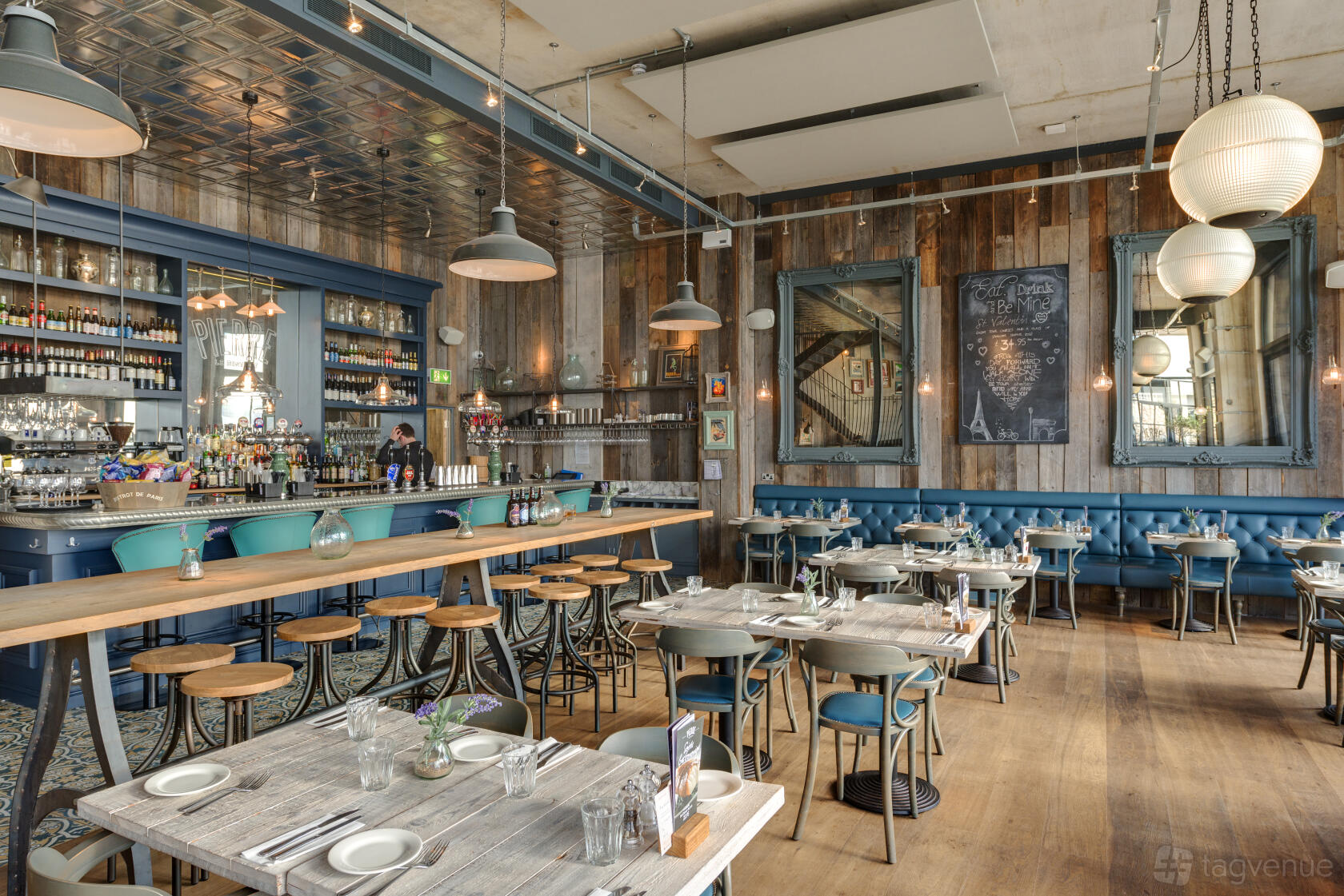 A bar in a restaurant with a long wooden counter, blue bar stools, tufted banquette seating, and large hanging globe lights at Bistrot Pierre Torquay.