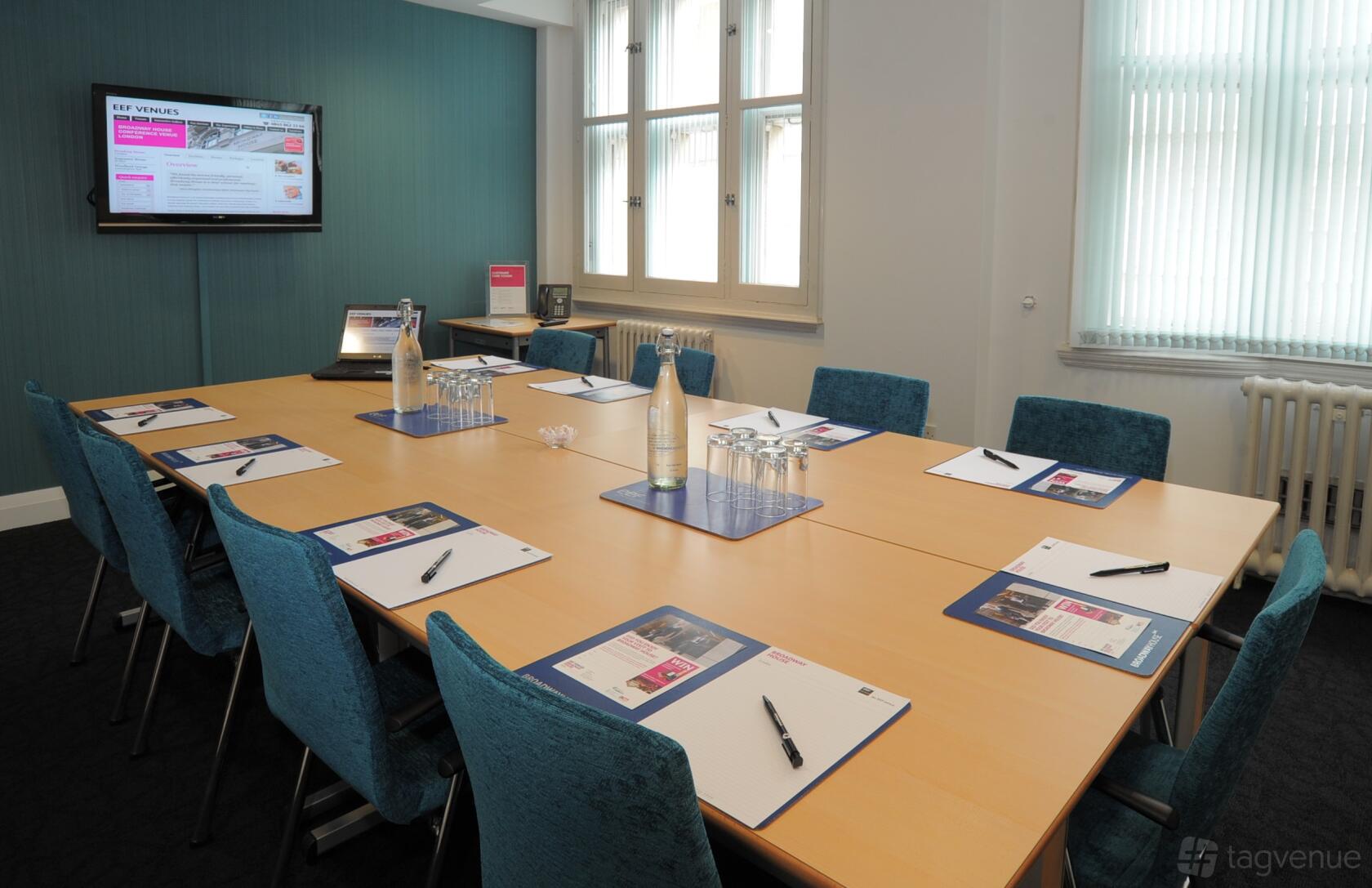A meeting room with a rectangular table, teal chairs, bottled water, and a wall-mounted screen at Broadway House.