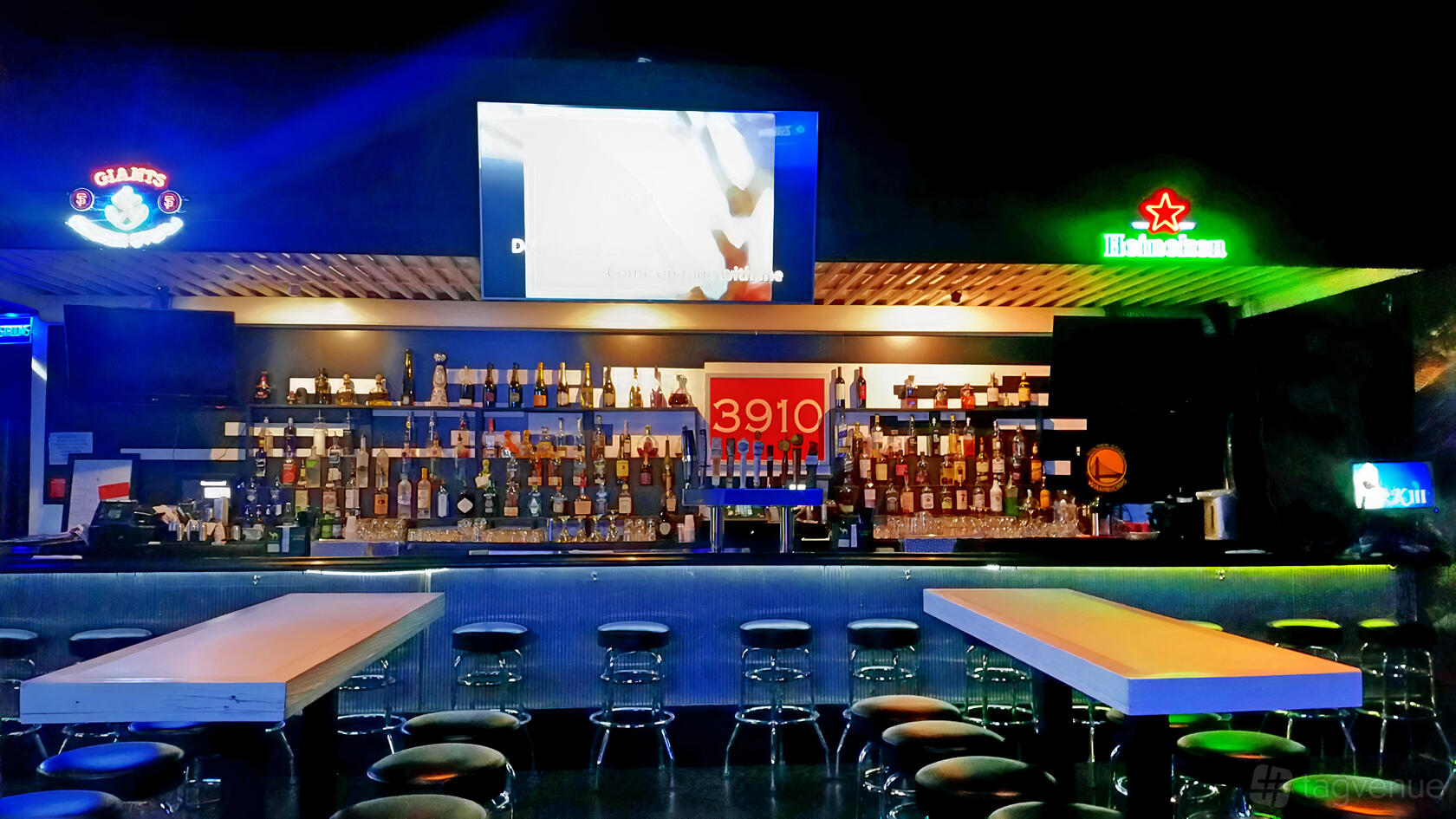 A bar with a long counter, backlit shelves stocked with liquor bottles, barstools, and neon signage at 3910 Lounge.