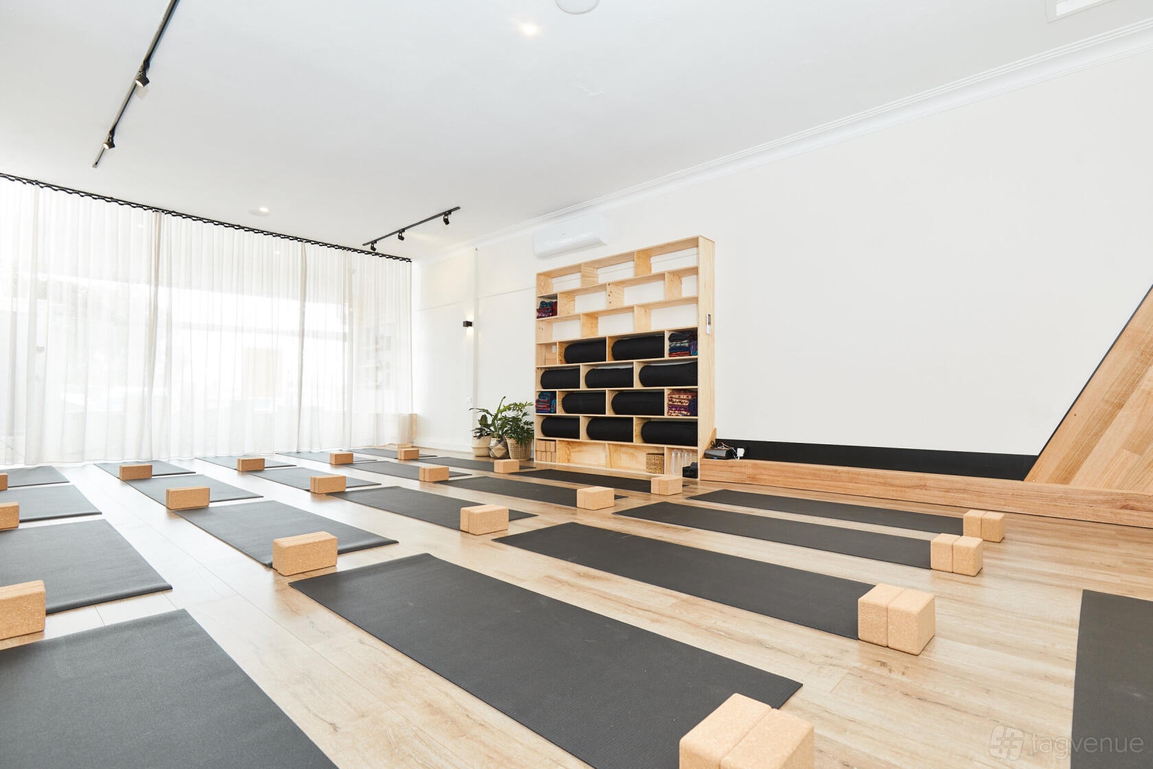 A yoga studio with light wood floors, yoga mats, cork blocks, and a shelving unit with rolled blankets at Oshun Yoga.