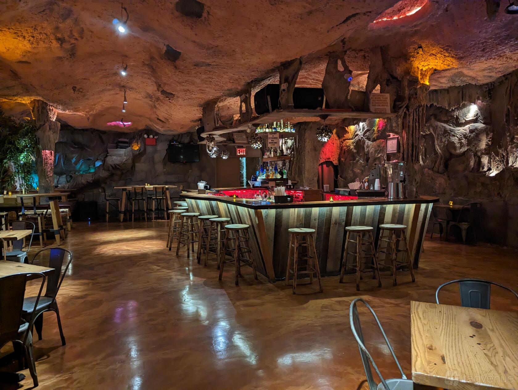 A bar in a restaurant with cave-like stone walls, rustic wooden stools, and glowing red bar lighting at La Caverna.