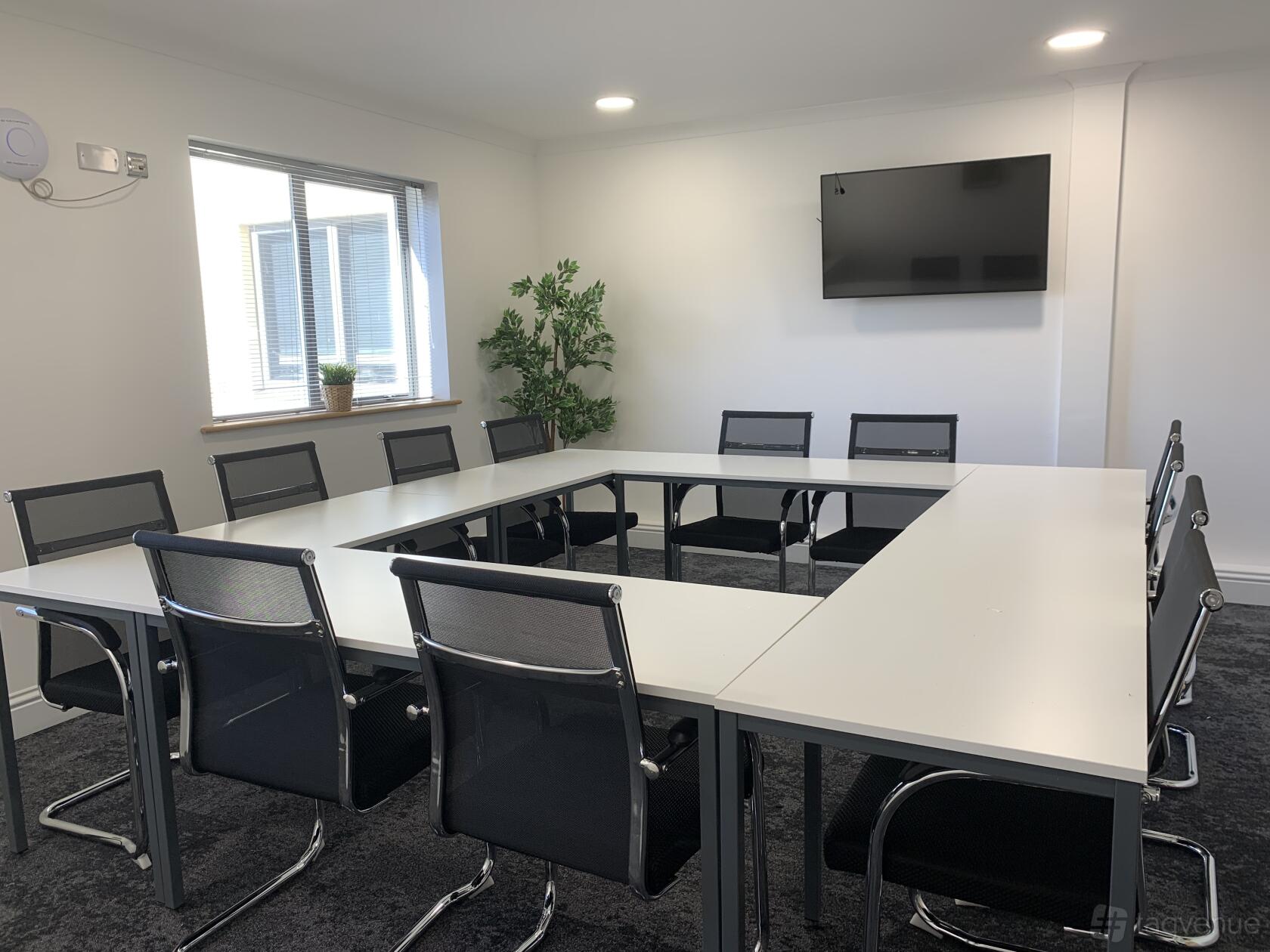 A meeting room with U-shaped white tables, black mesh chairs, a wall-mounted screen, and a window at Biz Hub City View House.