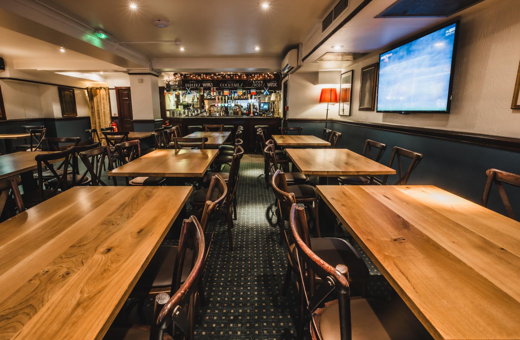 A pub function room with wooden tables, cushioned chairs, carpeted floors, and a stocked bar at The Hercules Pillars.