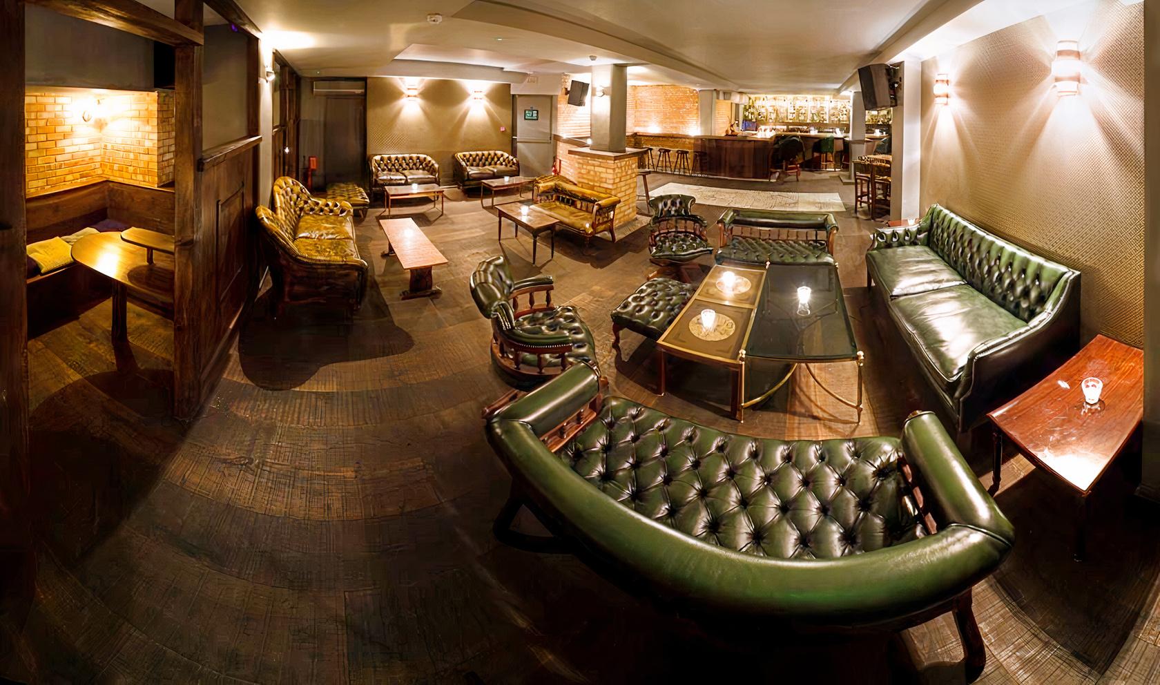 A cocktail bar with tufted leather sofas, wooden tables, and exposed brick accents at Basement Sate.