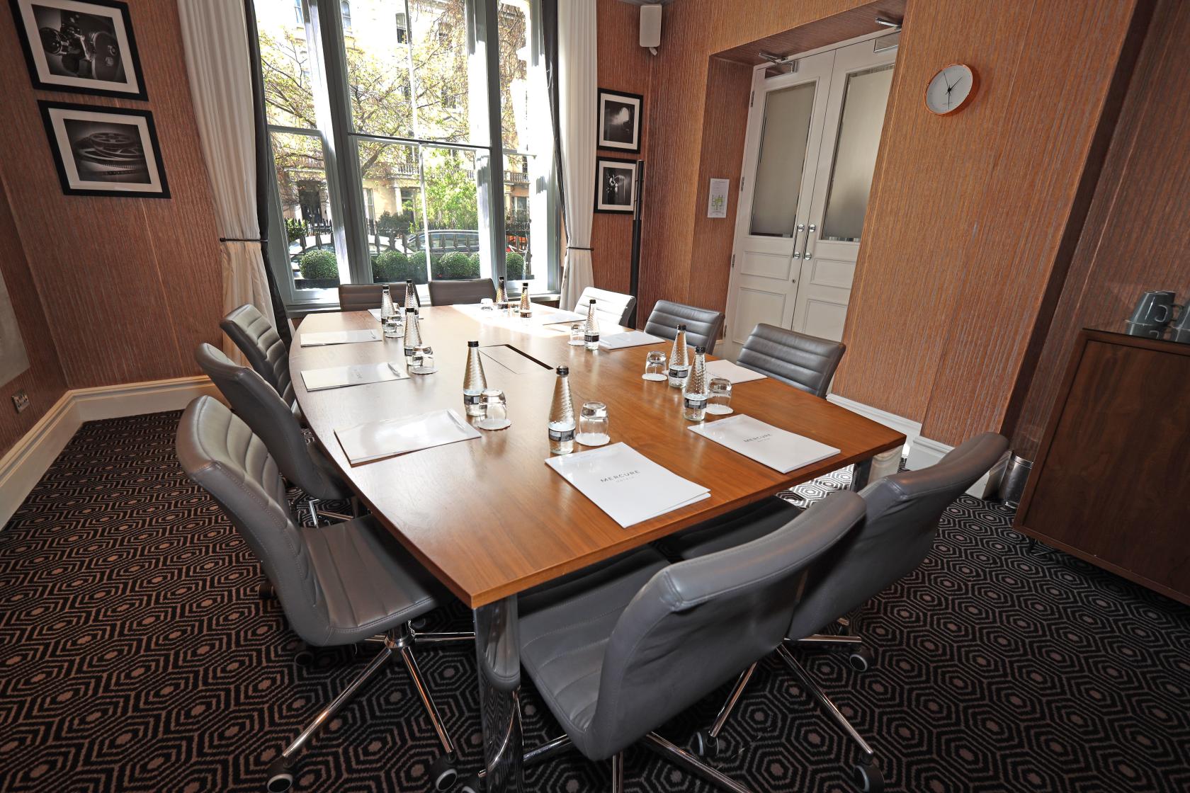 An event space in a hotel with floor-to-ceiling windows, a wooden meeting table, and grey chairs at Mercure London Hyde Park.