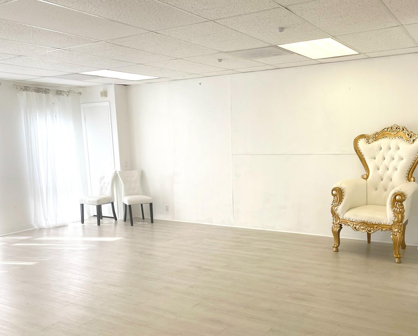 A photo studio with white walls, light wood floors, sheer curtains, and an ornate gold and white chair at West Houston Event & Studio Space.