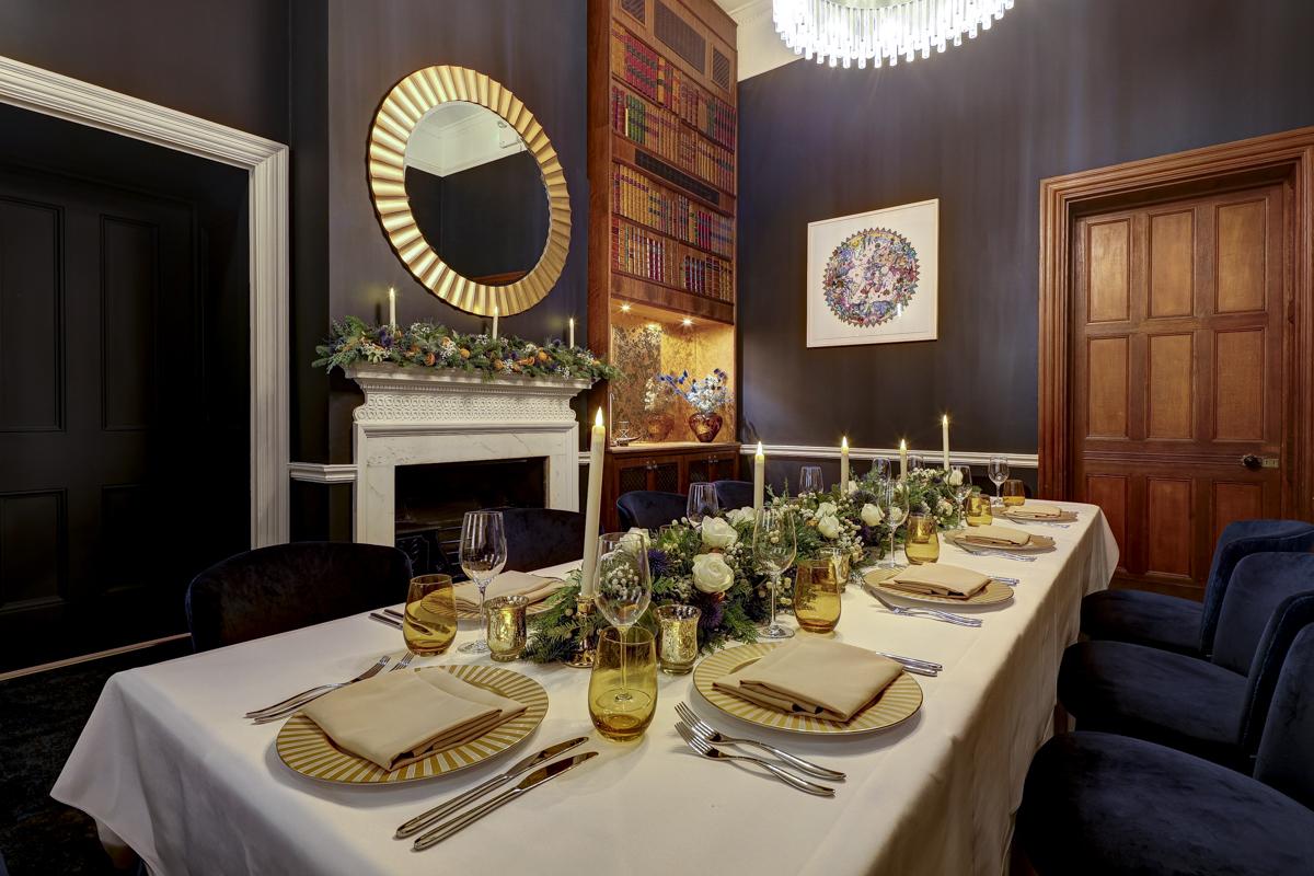 A dining room with a long banquet table, floral centerpiece, velvet chairs, and a decorative fireplace at Surveyors House at Rics Westminster.