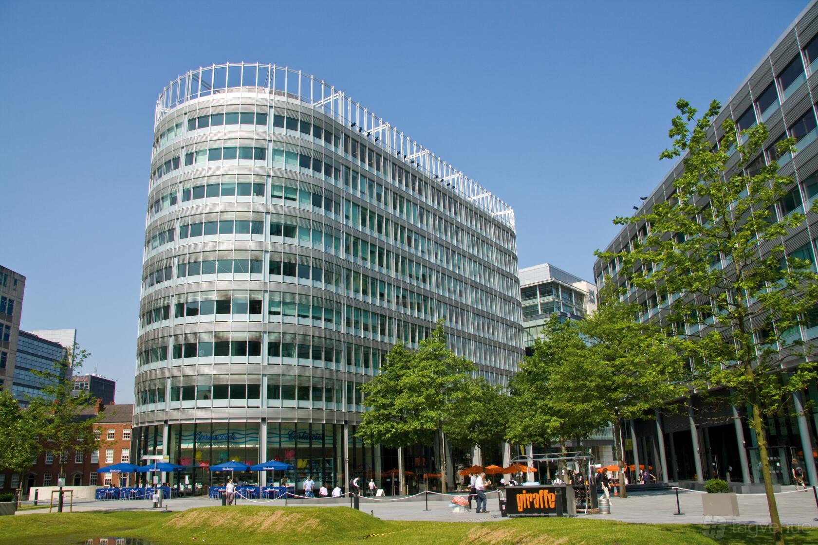 A meeting centre with a curved glass façade, outdoor seating, and trees at Landmark: Spinningfields, Manchester.