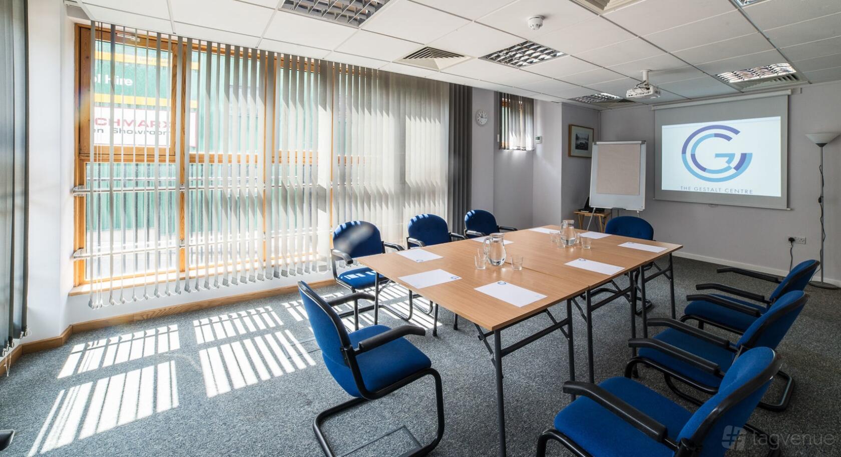 A meeting room with large windows, vertical blinds, a projection screen, and blue chairs at The Gestalt Centre in London.
