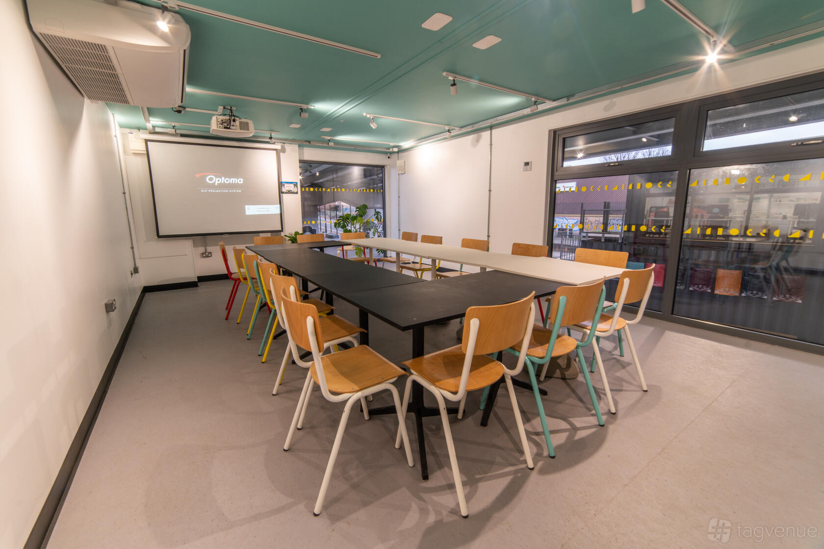 A meeting room with a U-shaped arrangement of wooden chairs and tables, projector, and large windows at Projekts Skatepark.