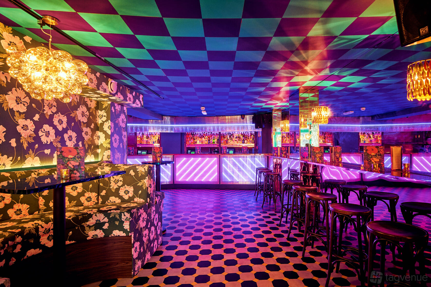 A bar with a checkered ceiling, patterned wallpaper, glowing neon-lit bar counter, and wooden stools at Blame Gloria Clapham Junction.