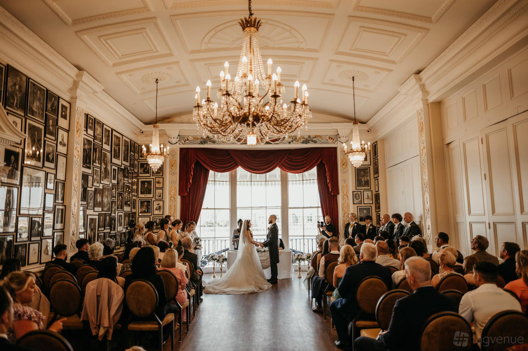 A dining room in a pub with chandeliers, framed portraits on the walls, and large windows with red curtains at Trafalgar Tavern.