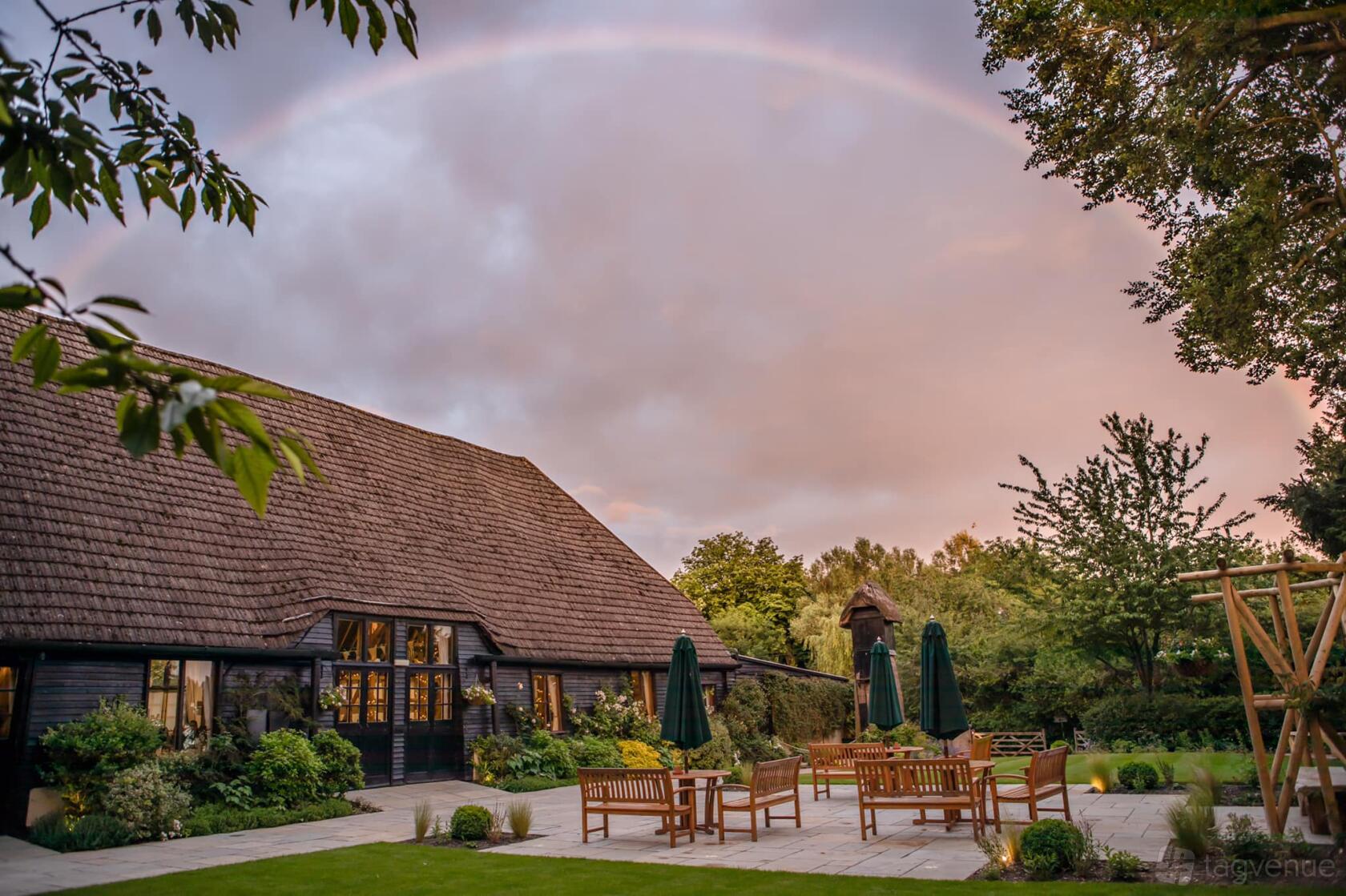 A rustic barn venue with a stone patio, wooden outdoor furniture, and surrounding gardens at Clock Barn.