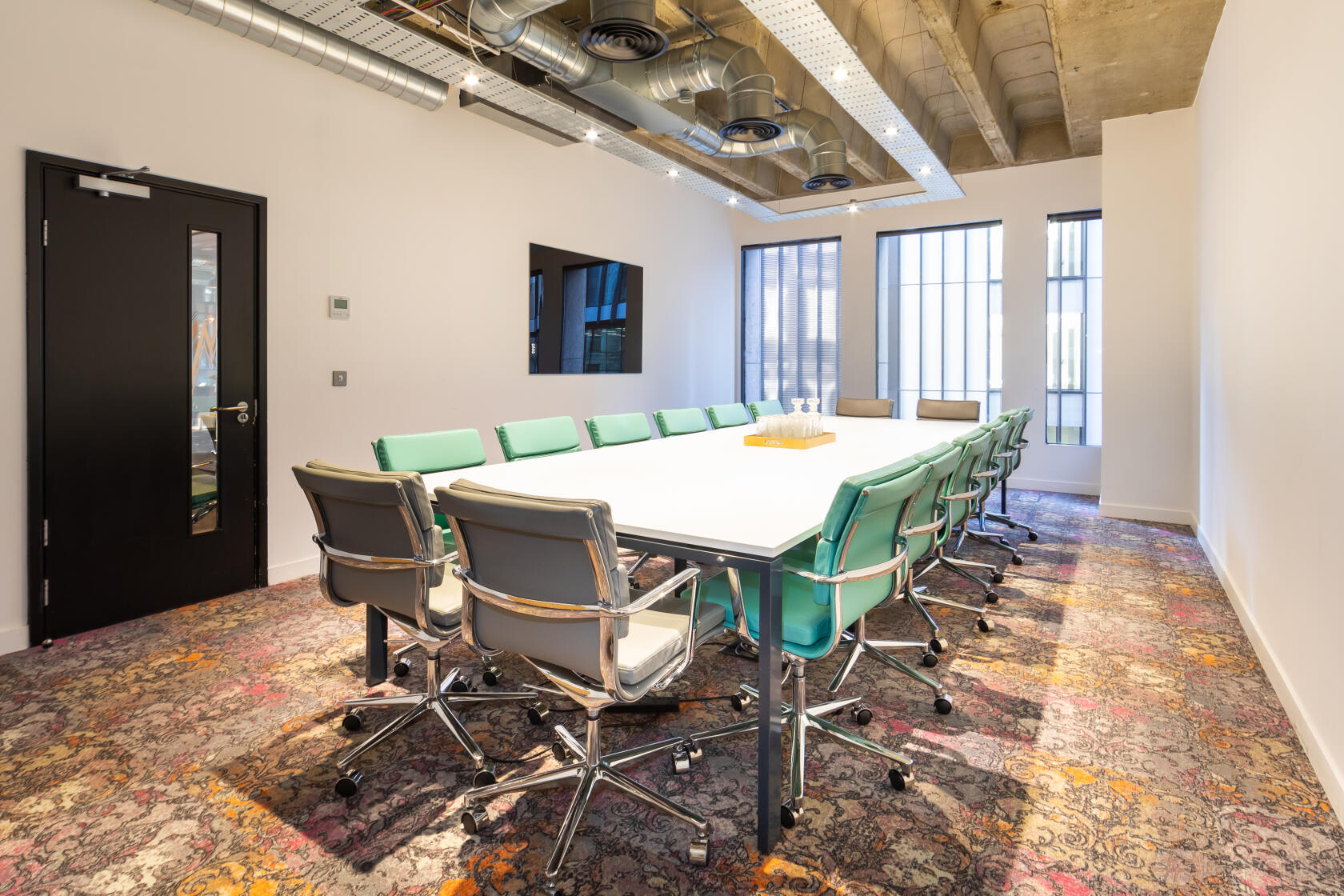 A meeting room with a long white conference table, green chairs, large windows, and exposed ductwork at The Space Aldgate.