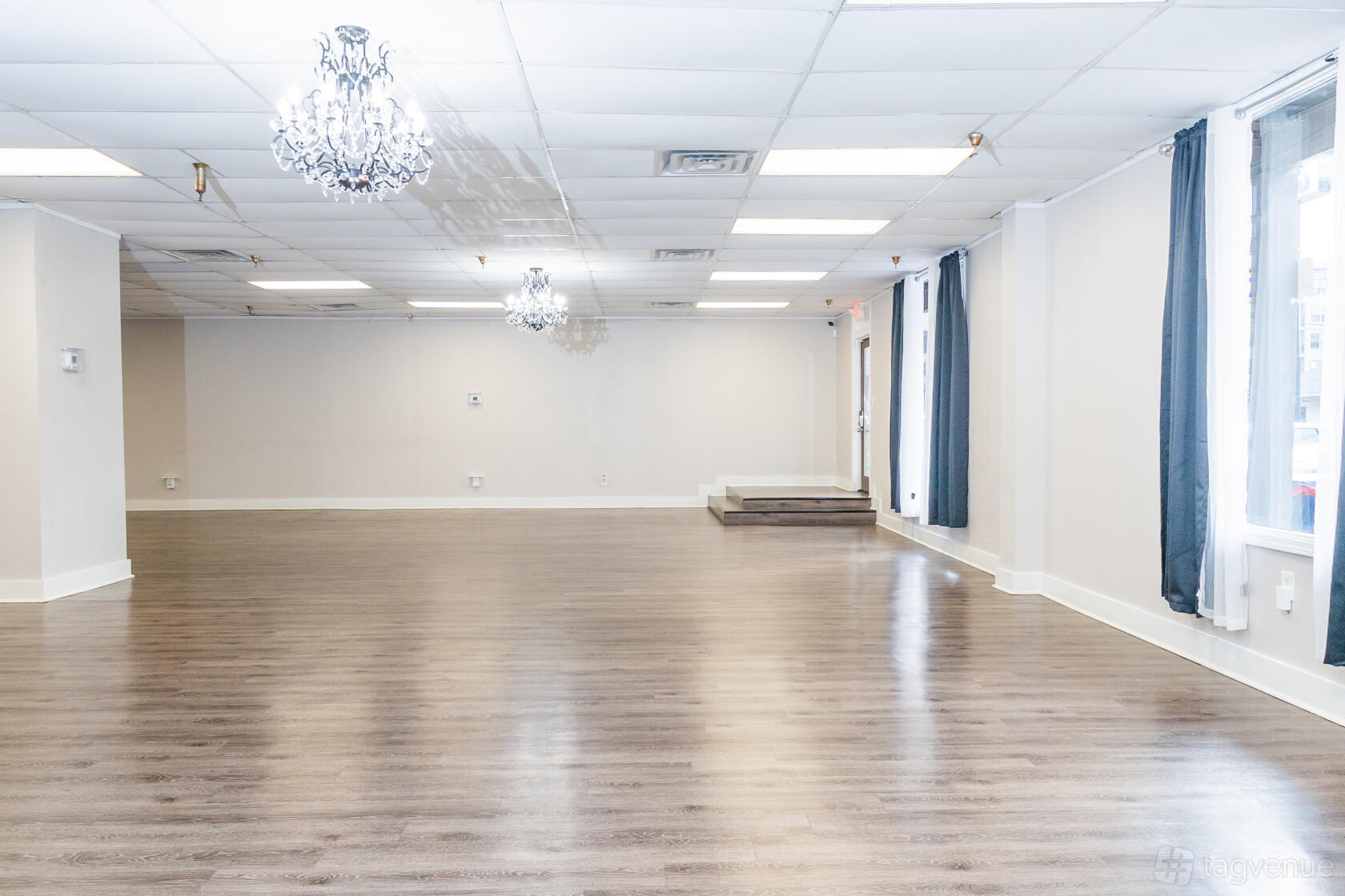 An event venue with wood flooring, white walls, chandeliers, and large windows with black curtains at Space in Time.