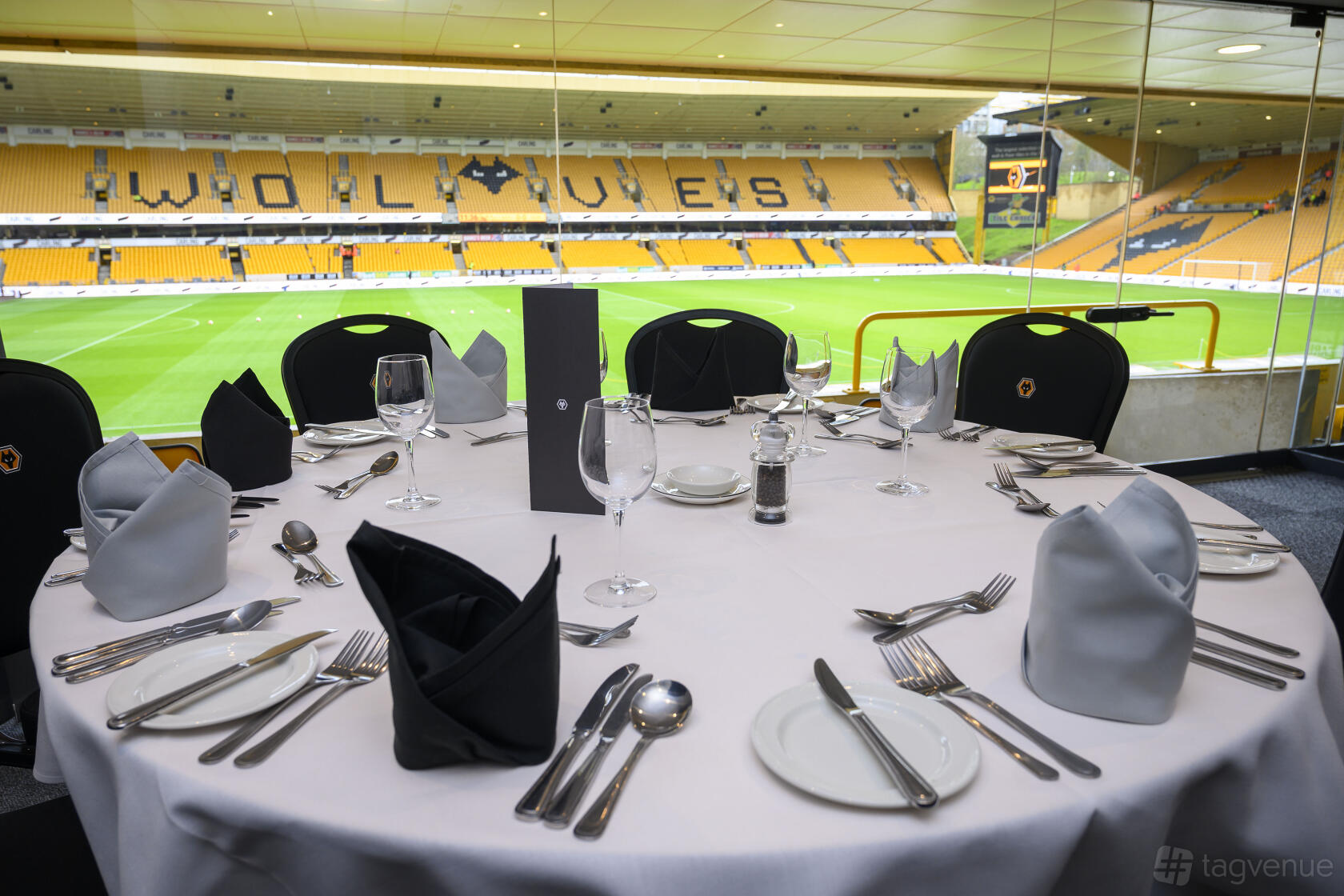A stadium suite with floor-to-ceiling windows, round table set with glassware and napkins overlooking the pitch at Molineux Stadium.