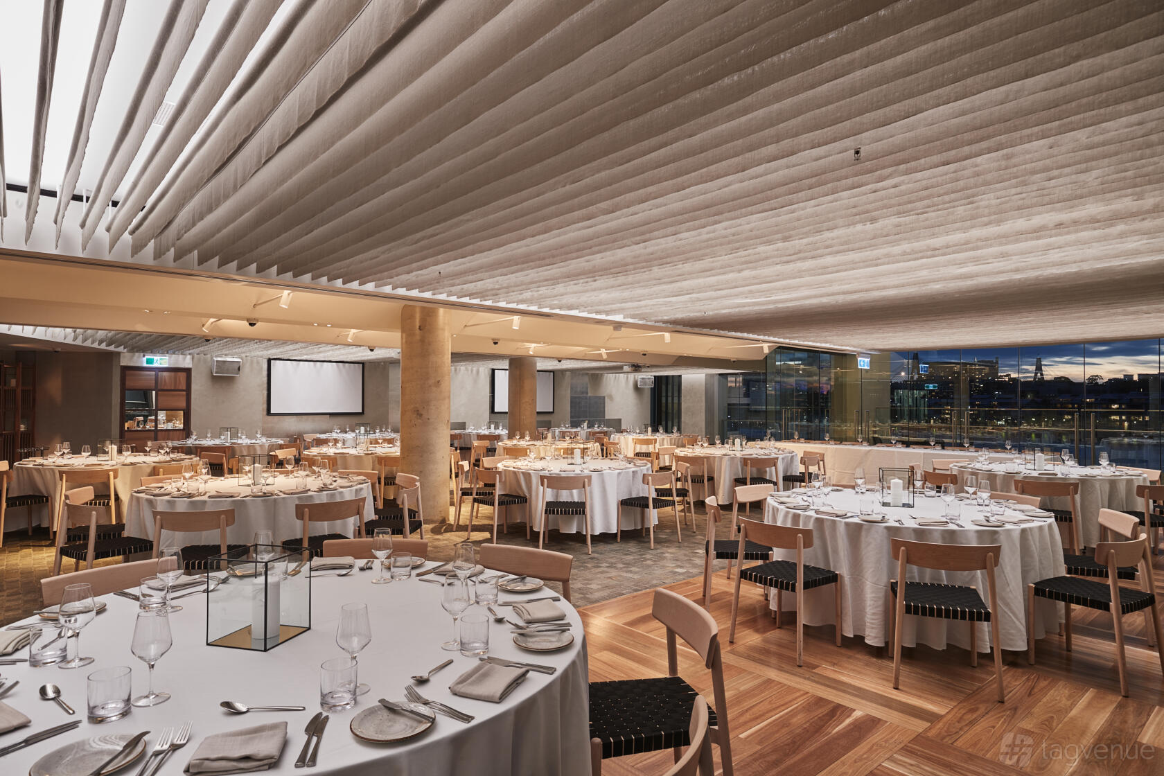 A banquet hall with round tables draped in white linens, wooden chairs, and large windows at 12-Micron.