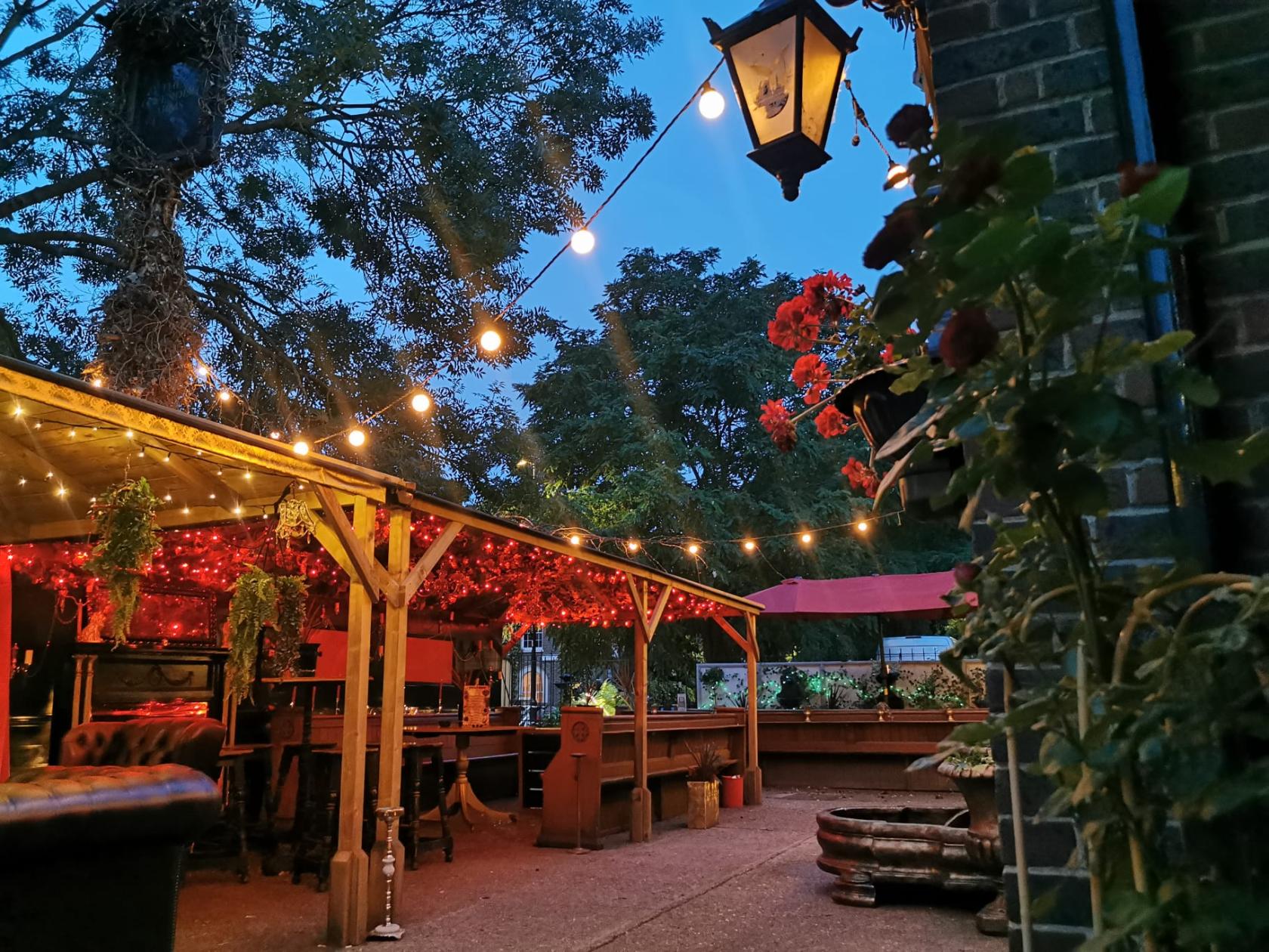 An outdoor pub garden with string lights, hanging plants, and cushioned seating at The Cavendish Arms.