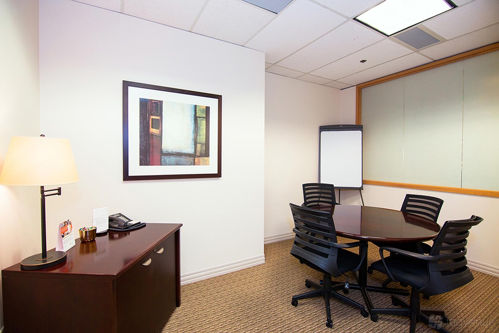 A meeting room with a round table, four black chairs, a flip chart, and framed wall art at Premier Workspaces - Eastside Office Center.