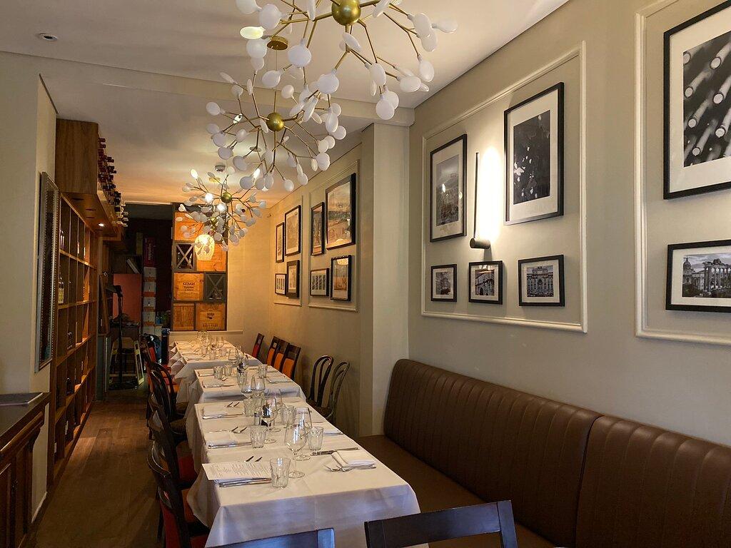 A dining room with long tables covered in white linens, padded bench seating, and framed photos on the wall at Bacco Ristorante Italiano.