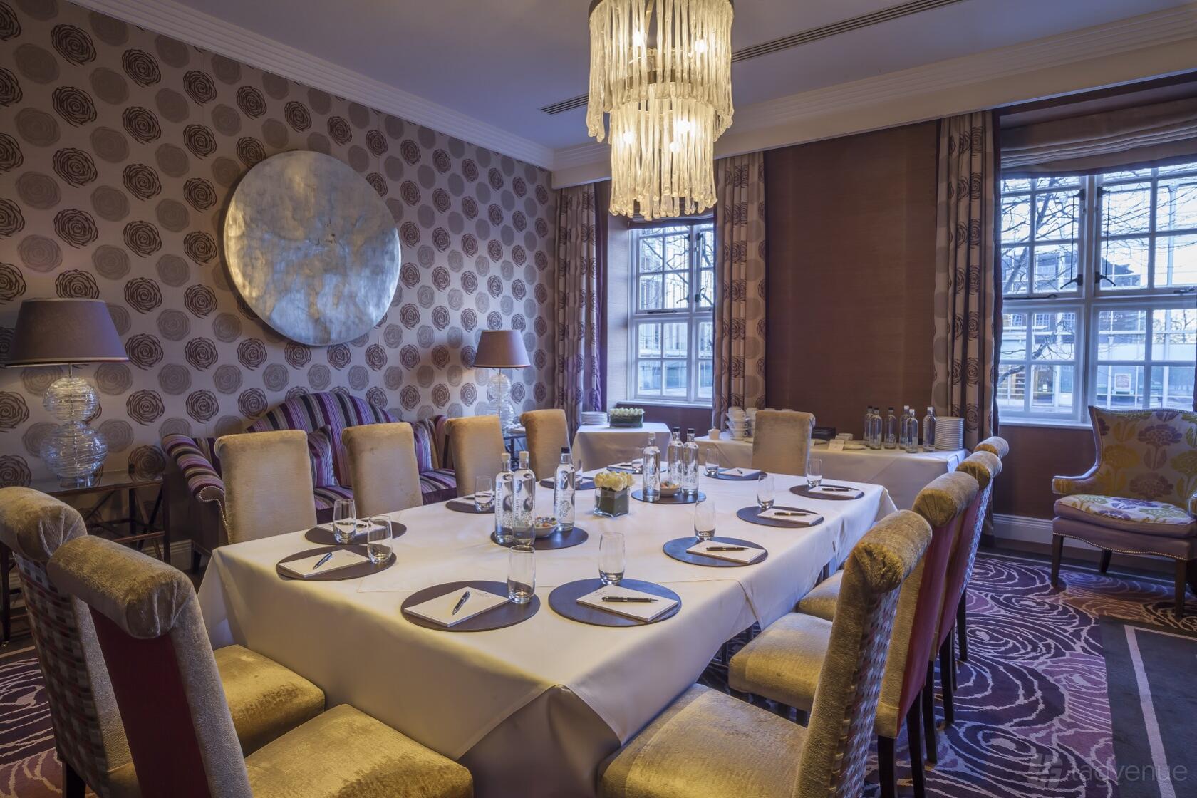 A hotel meeting room with a rectangular table, plush chairs, patterned wallpaper, and a chandelier at The Roseate Reading.