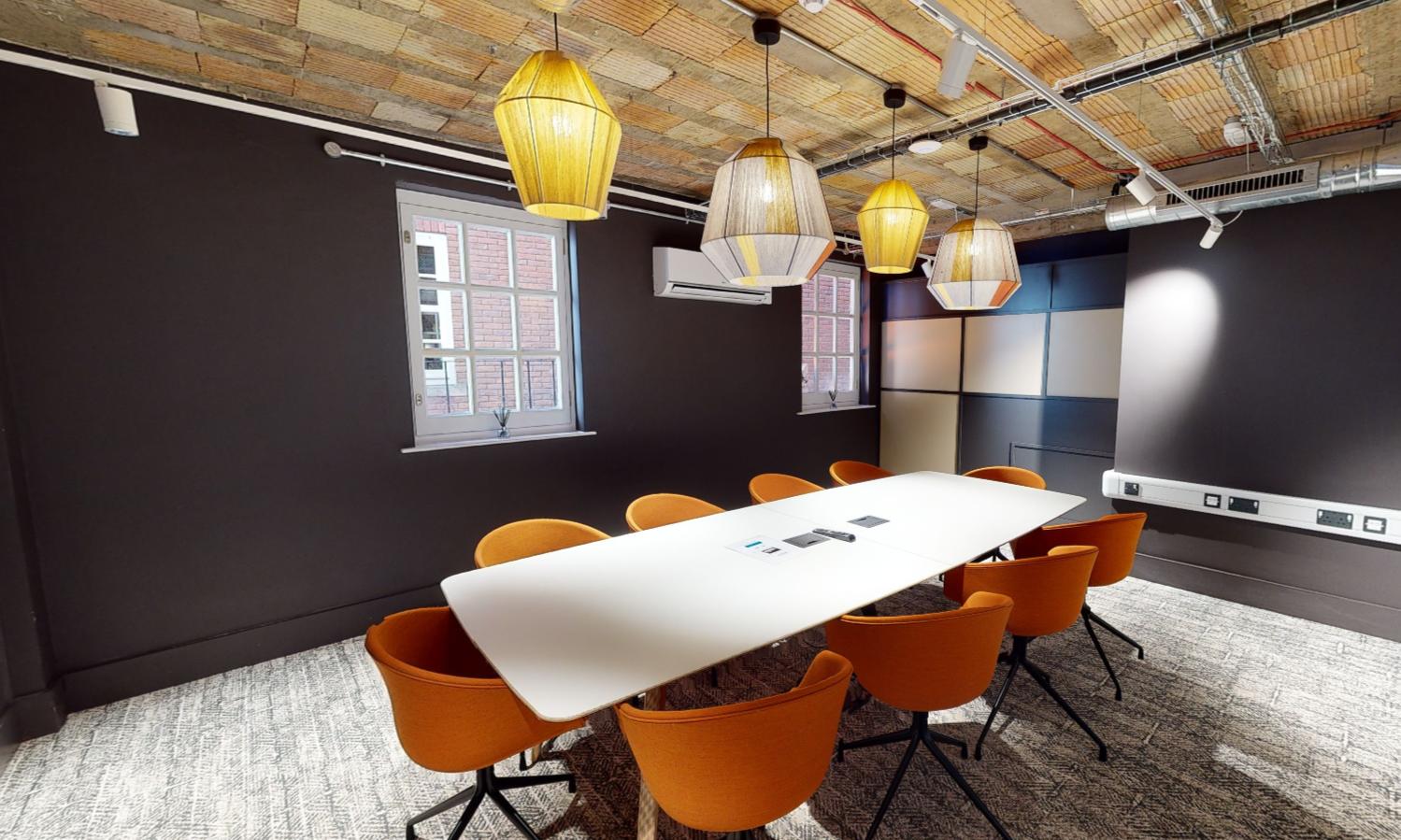 A meeting room with a long white table, orange chairs, exposed brick ceiling, and large pendant lights at The Boutique Workplace Company.