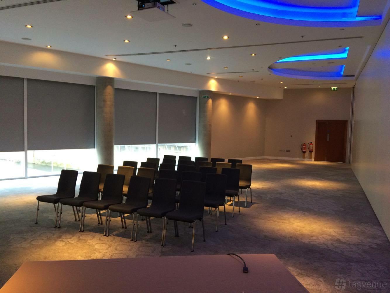 A ballroom with rows of black chairs, carpeted flooring, large covered windows, and uplighting at The Lowry Hotel.