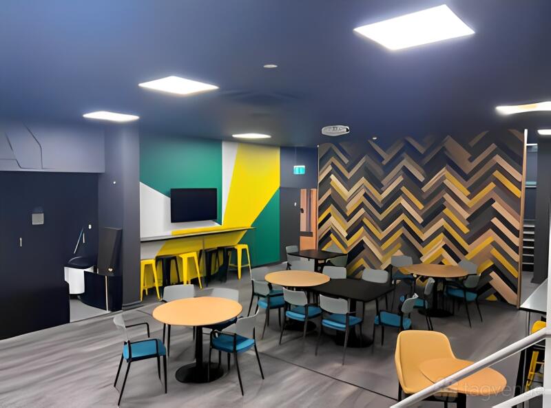 A stadium lounge with round tables, chevron accent wall, and yellow bar stools at Northampton Saints Stadium.