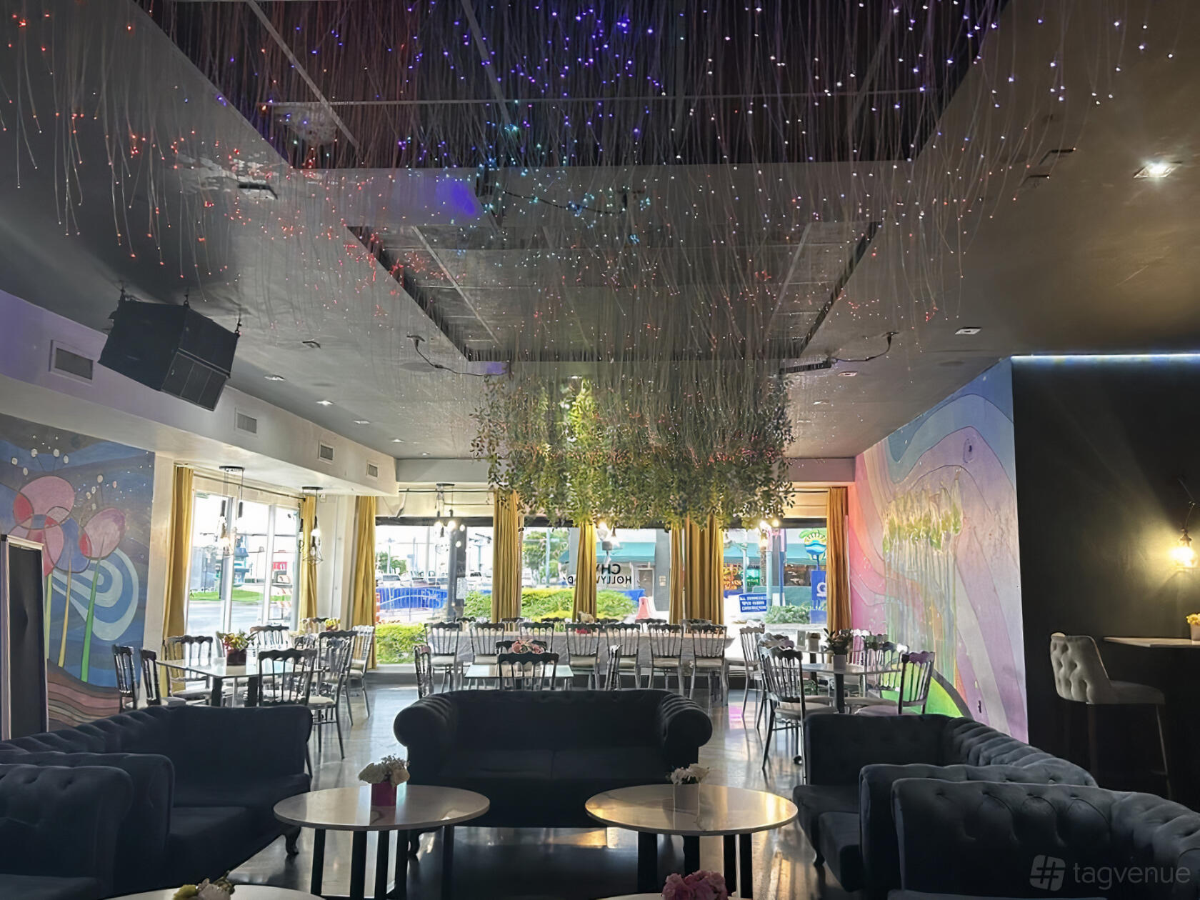 A restaurant with floor-to-ceiling windows, velvet sofas, round tables, and hanging decorative lights at Chwa Hollywood.