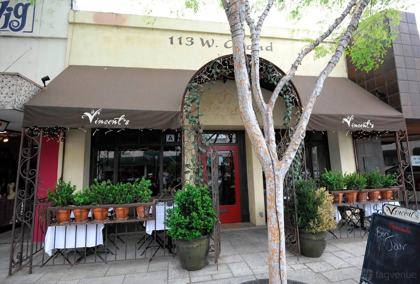 A pub with a brown awning, red door, potted plants, and sidewalk seating at Hunsakers.