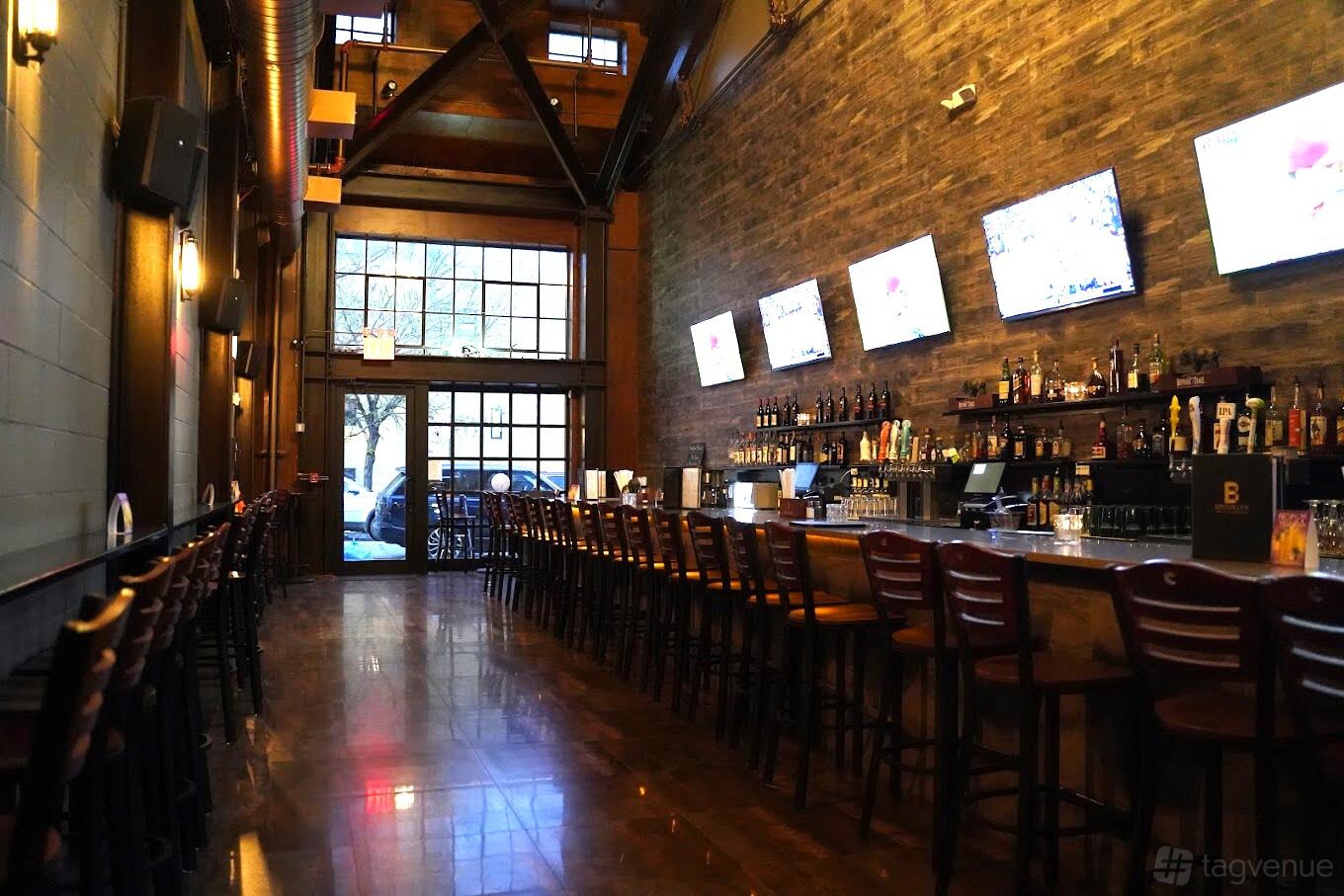 A bar with a long wooden counter, high-top chairs, wall-mounted TVs, and exposed brick walls at Brooklyn Billiards.