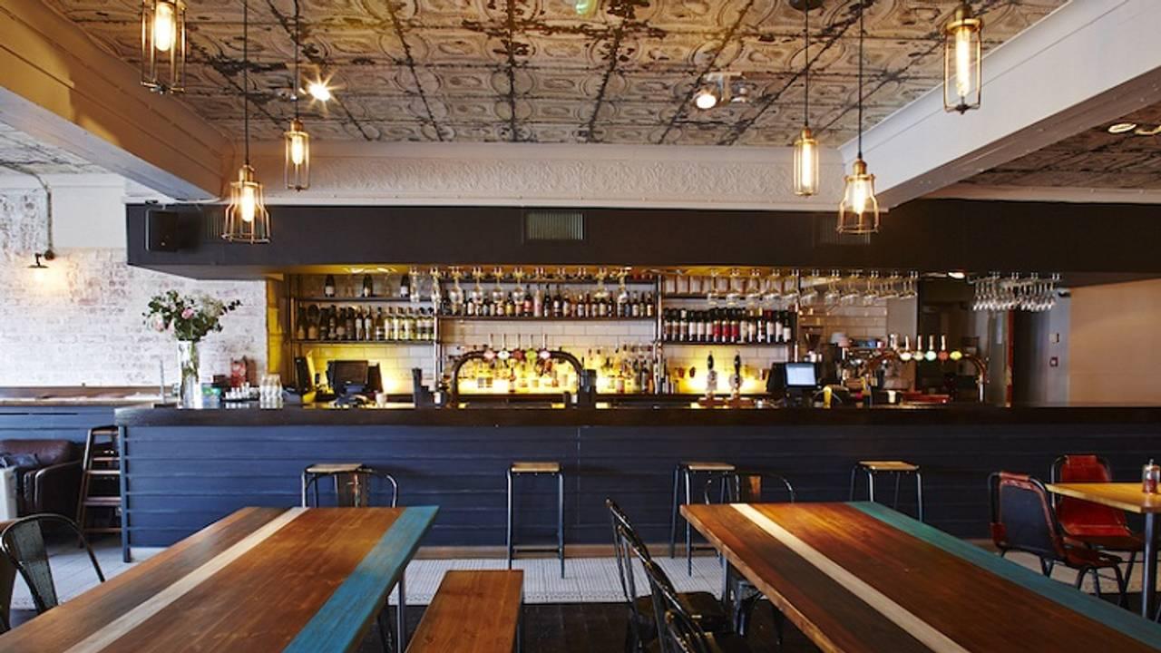 A dining room with long wooden tables, patterned tin ceiling, and hanging pendant lights at Luppolo Pizzeria, Wanstead.