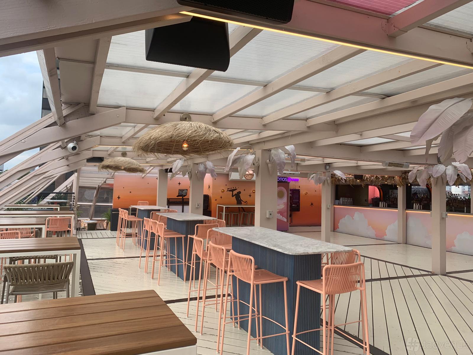 A rooftop bar with pastel decor, woven pendant lights, marble-topped high tables, and pink stools at Circe's Rooftop Waterloo.