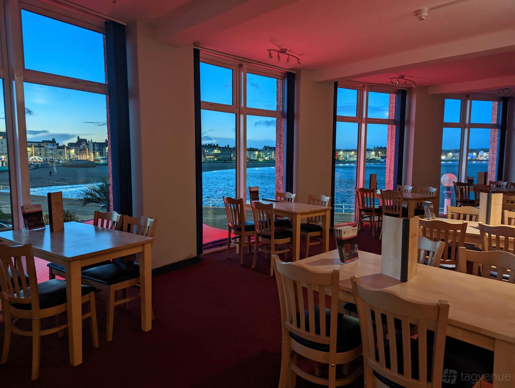 A piano room with large windows overlooking the waterfront and light wood tables at Weymouth Pavilion.