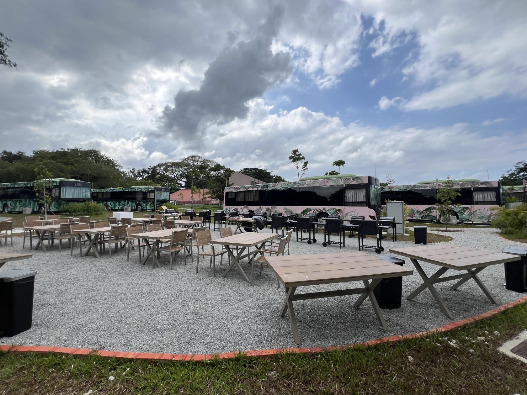 An outdoor venue with wooden tables, chairs, and gravel flooring surrounded by greenery at The Bus Collective.
