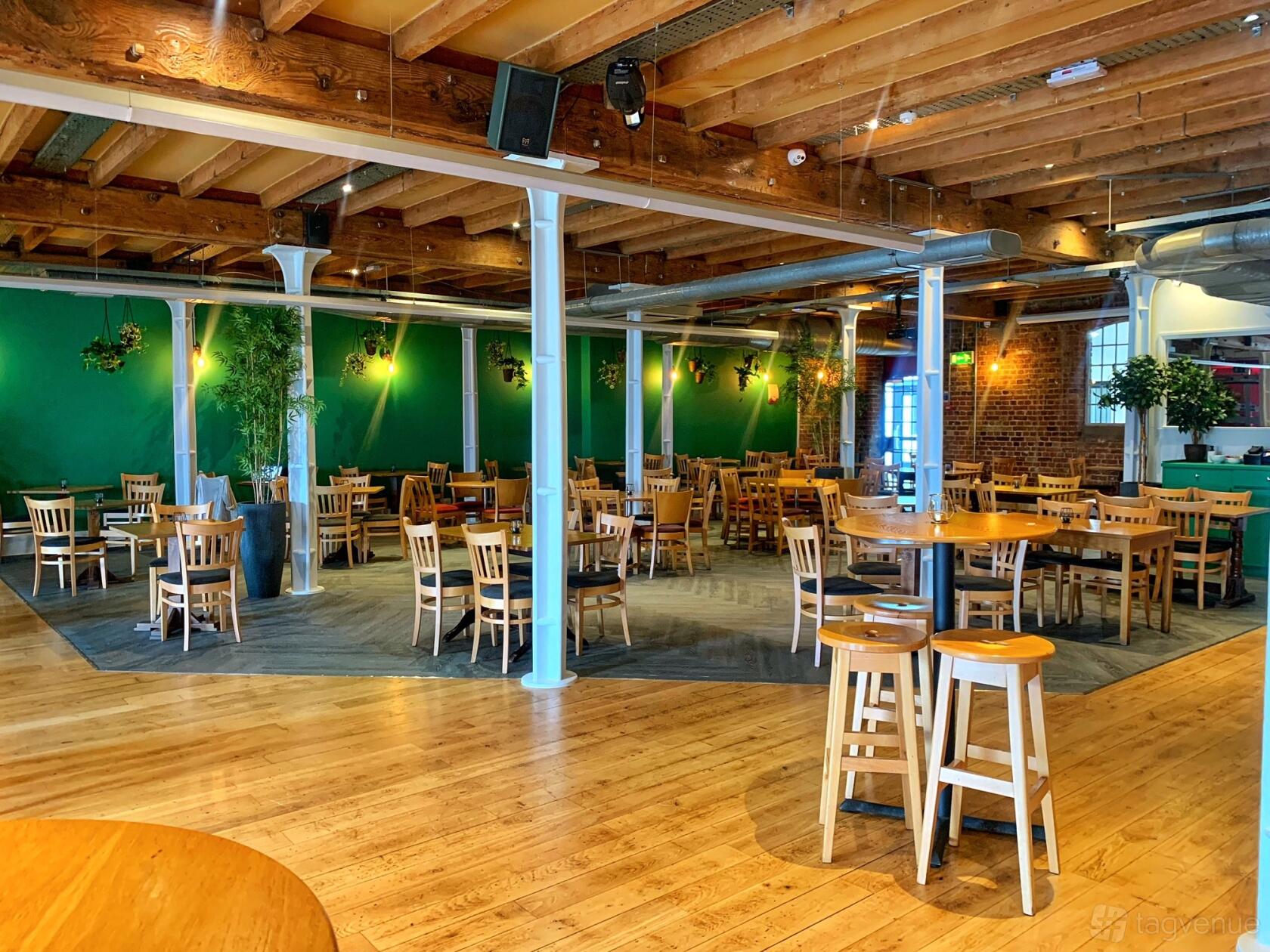 An event space with wooden tables and chairs, exposed beams, and hanging plants at Fox@ExCeL.