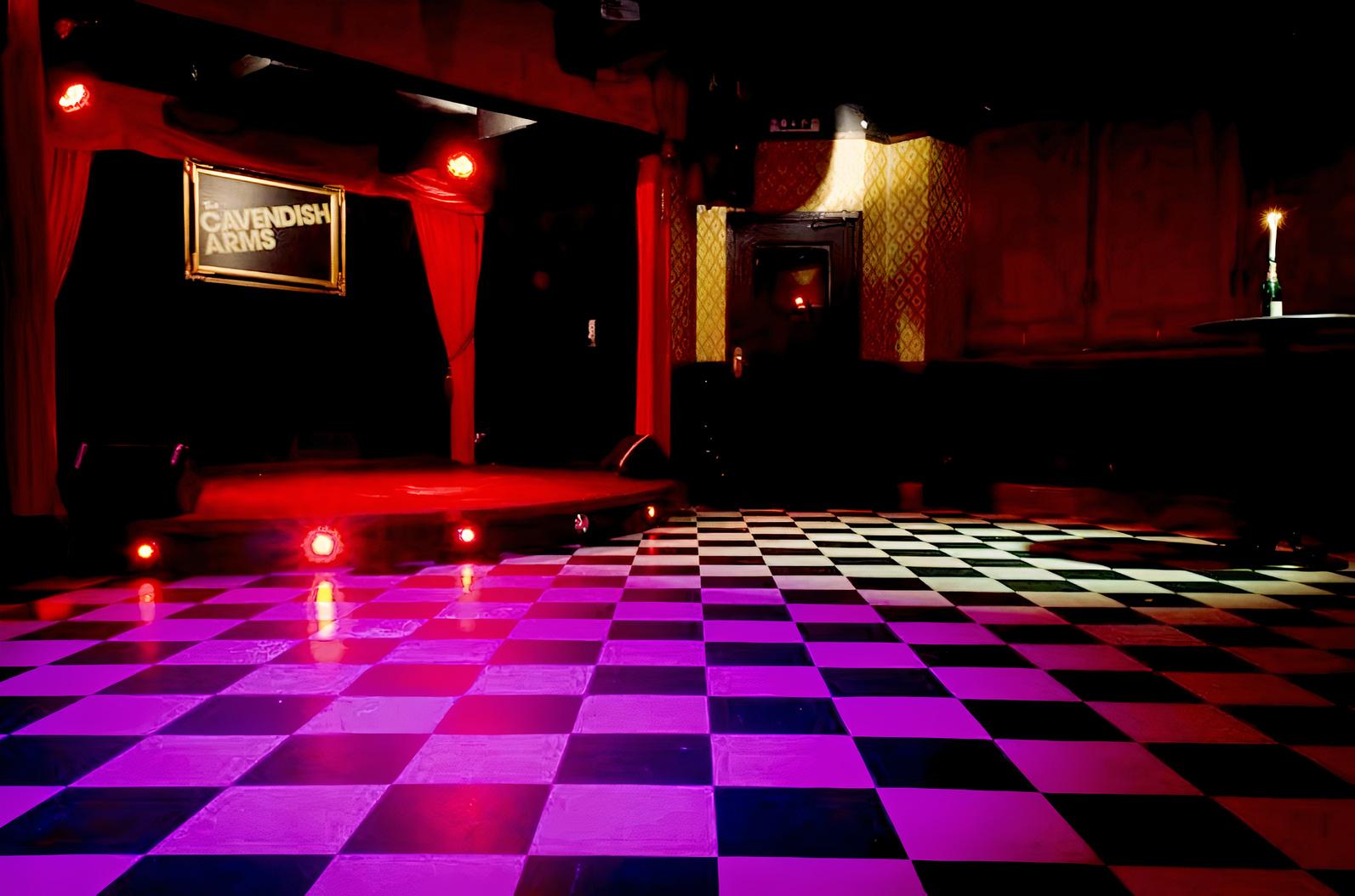 A function room in a pub with a black and white checkered floor, small stage, and red curtains at The Cavendish Arms.