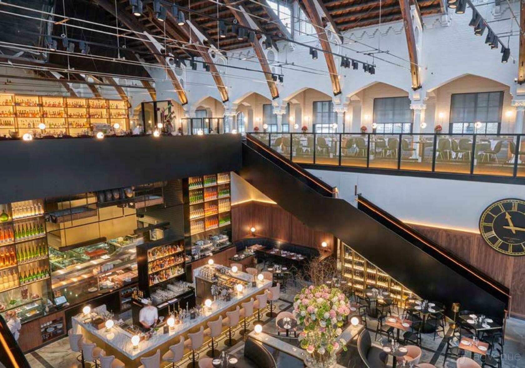 A restaurant with high vaulted ceilings, mezzanine seating, and a central open kitchen at German Gymnasium.