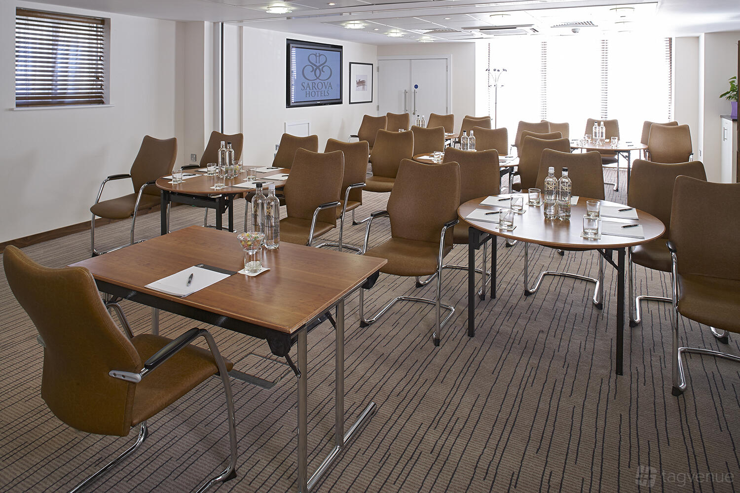 A function room with round tables, brown chairs, bottled water, and notepads in the Buckingham Suite at Sir Christopher Wren Hotel.