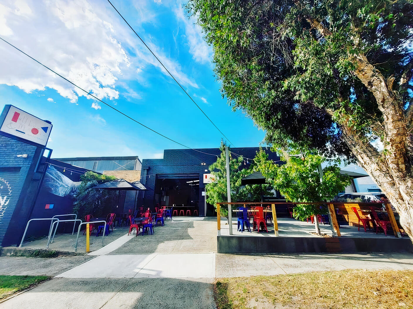 An outdoor bar patio with colorful chairs, picnic tables, and large trees at Tallboy and Moose.
