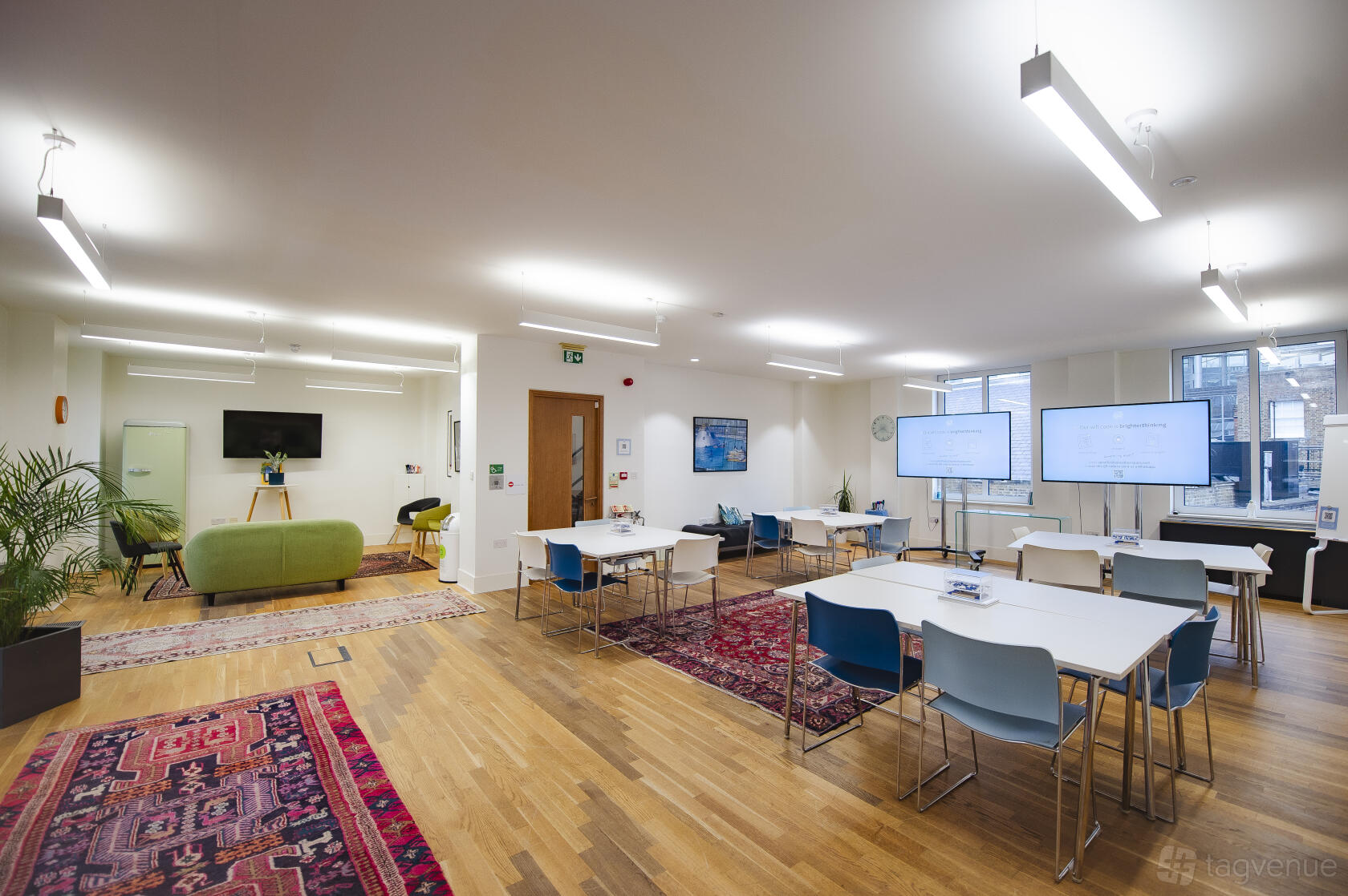 An event space with light wood floors, colorful rugs, modular tables, blue chairs, and large screens at Wallacespace Spitalfields.