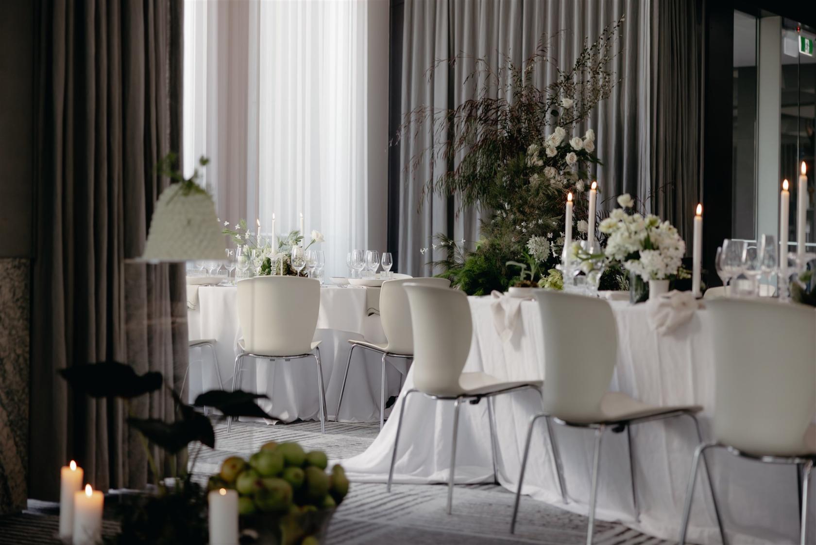 A hall with white chairs, draped tables, tall candles, floral centerpieces, and floor-to-ceiling curtains at Zinc at Fed Square.