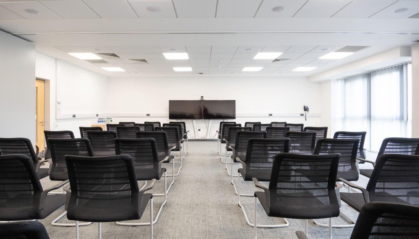 A classroom with rows of black mesh chairs facing a large wall-mounted screen and white walls at Rothamsted Conference Centre.