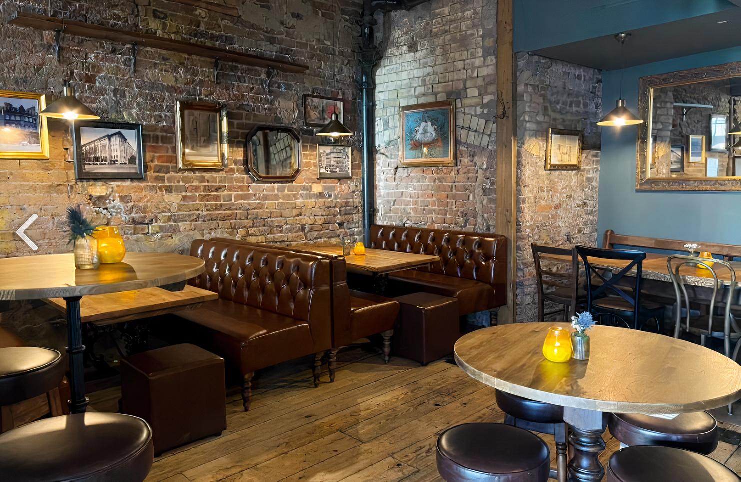 A pub with exposed brick walls, brown leather banquette seating, framed artwork, and wooden tables at The Clerkenwell Tavern.