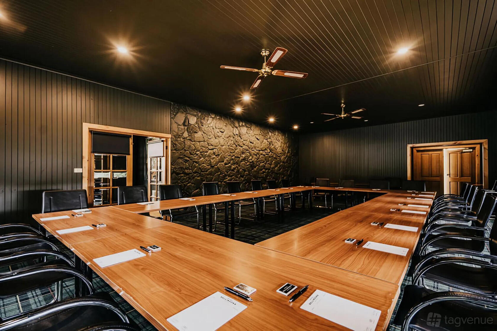 A hotel meeting room with U-shaped wooden tables, black chairs, notepads, and a stone accent wall at The Amalfi.