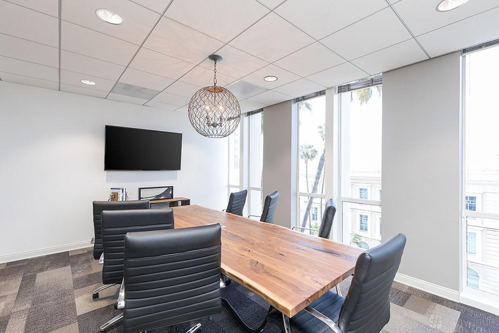 A meeting room with a wooden conference table, black leather chairs, flat-screen TV, and floor-to-ceiling windows at Premier Workspaces - Beverly Hills.