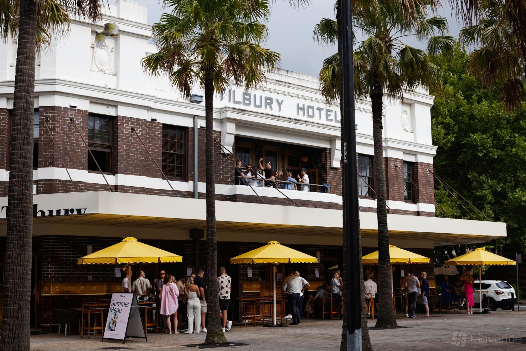 A pub with an outdoor bar area, brick facade, yellow umbrellas, and palm trees at The Tilbury Hotel.
