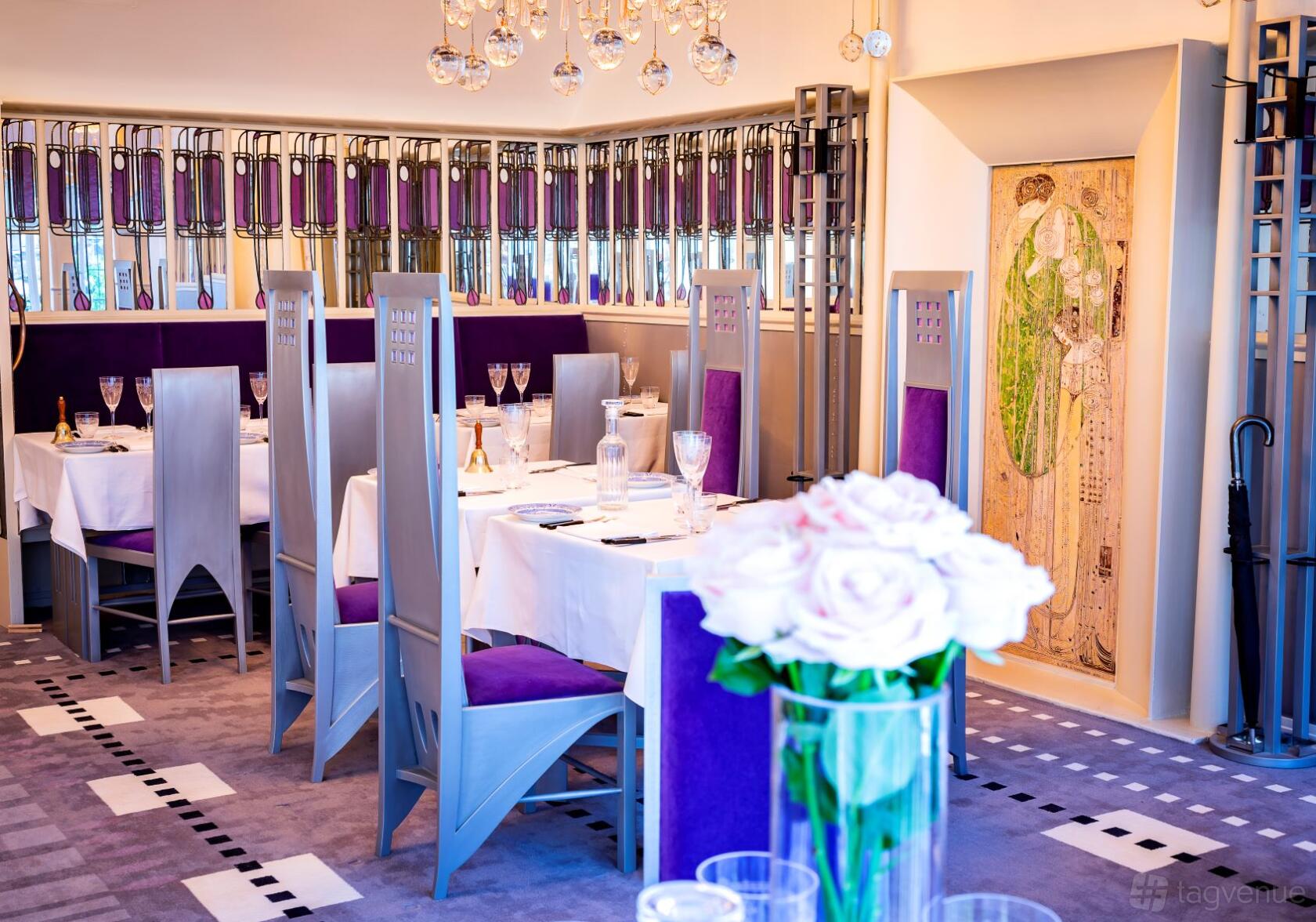 A dining room with purple upholstered chairs, stained glass accents, and white tablecloths at Mackintosh at the Willow.