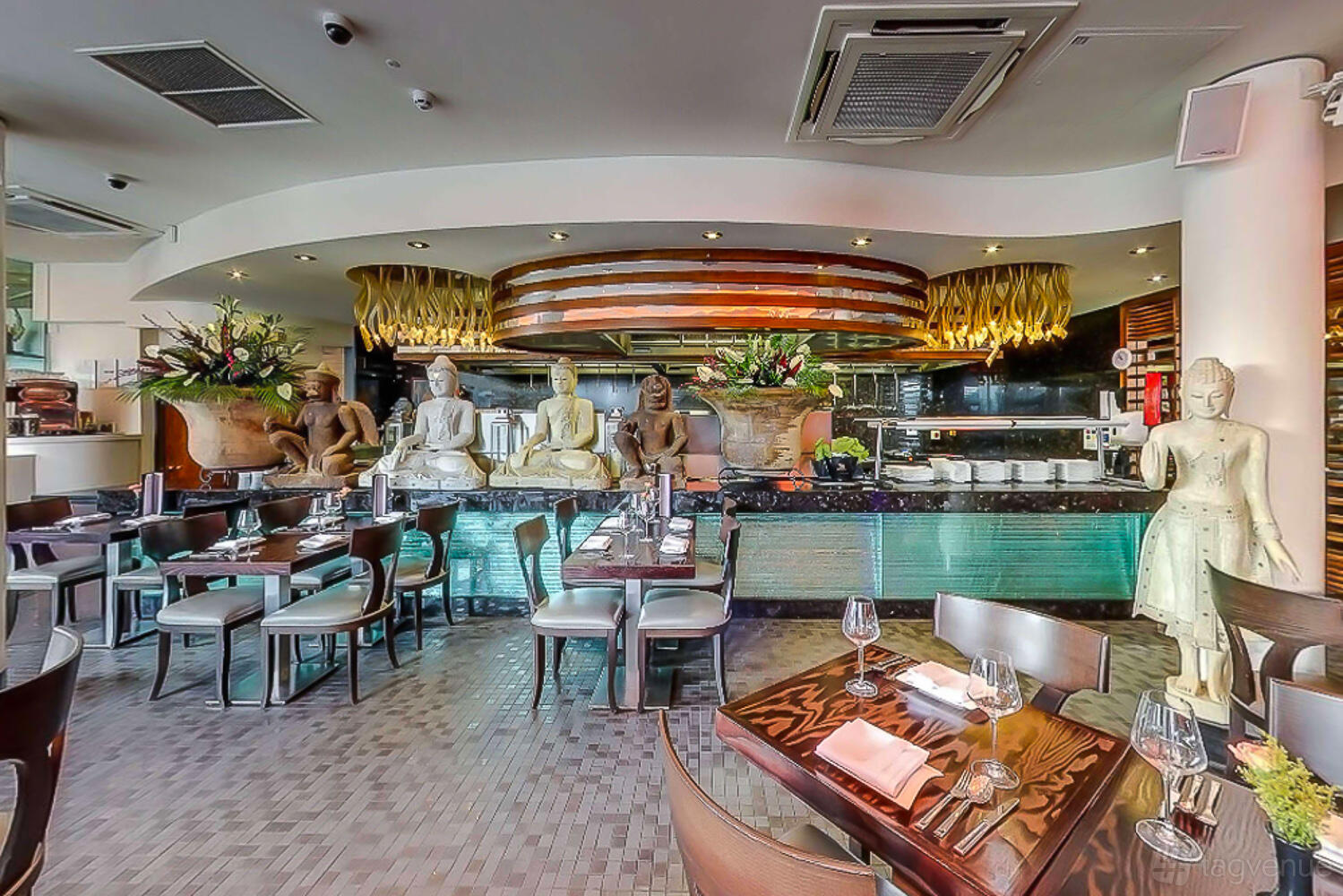 A restaurant with carved statues, open kitchen, and tables set with glassware at Chaophraya Birmingham.